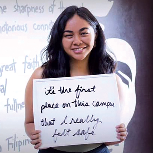 A young woman smiles holding a white board reading It's the first place on this campus that I really felt safe