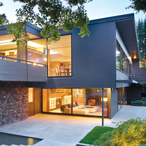 A large grey house in the midcentury style with warm light inside and a manicured yard outside.