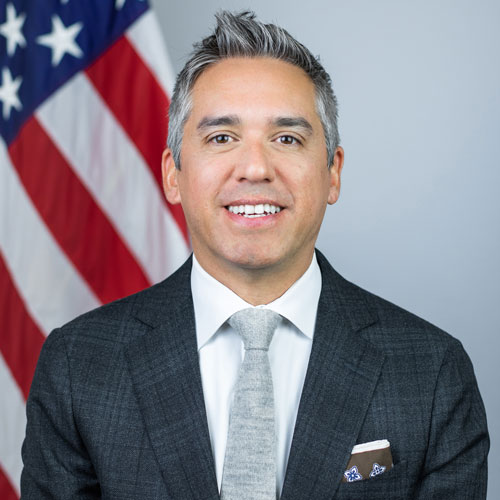Headshot of Cristobal Alex wearing a suit with the American flag in the background