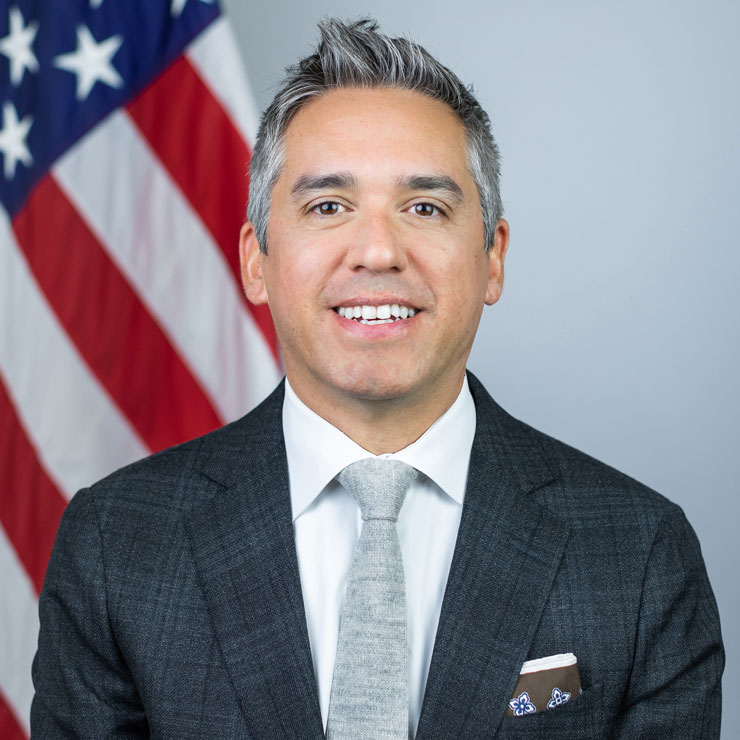 Headshot of Cristobal Alex wearing a suit with the American flag in the background