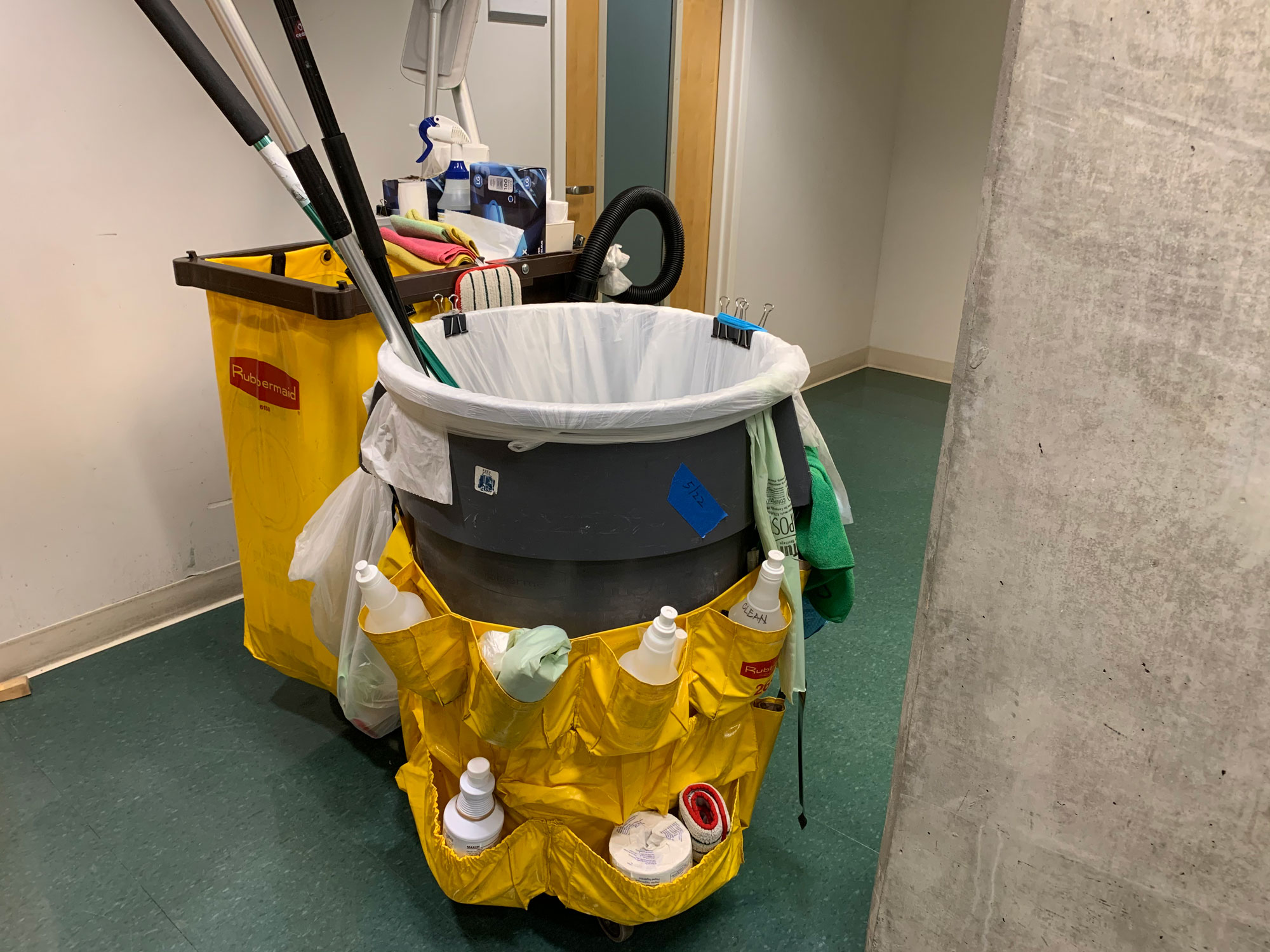 A trash can with a cleaning supplies cart in an office hallway.