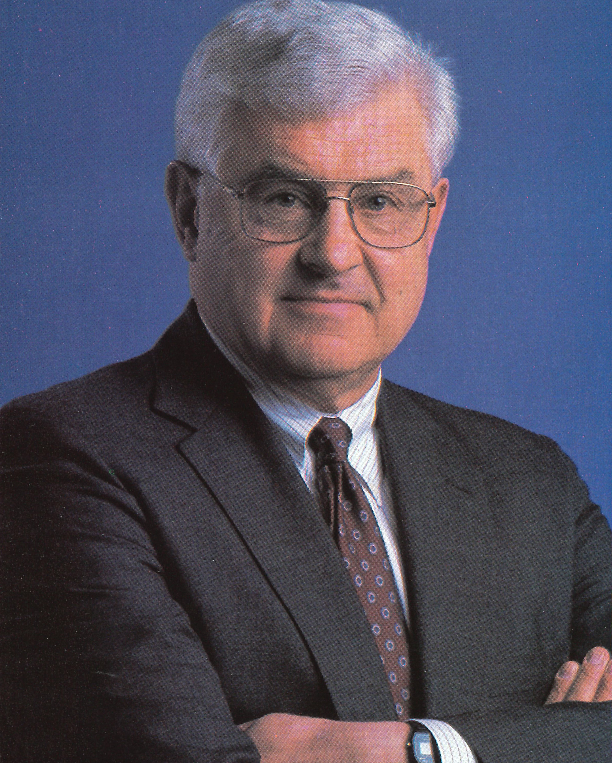 Headshot of Tracy King, a white-haired man in a grey suit with his arms crossed