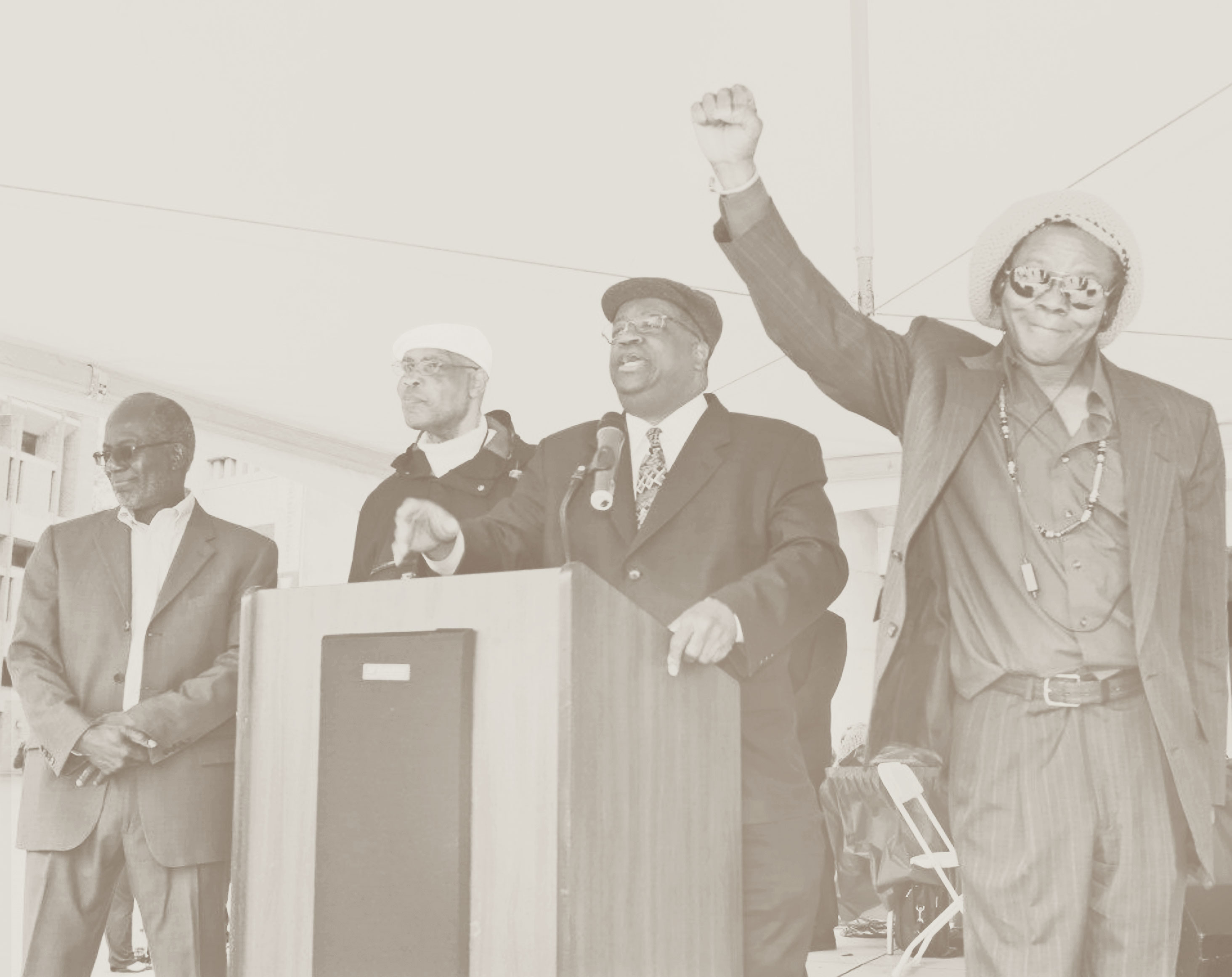 Four men stand near a podium; the man on the far right is holding a Black power fist