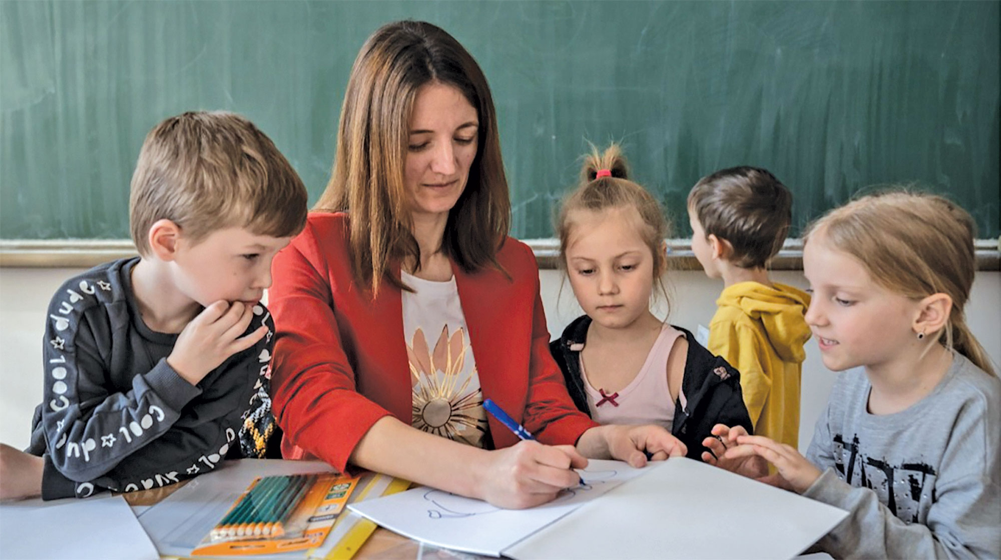 A woman wearing a red blazer sits at a table with four young children teaching a lesson on paper
