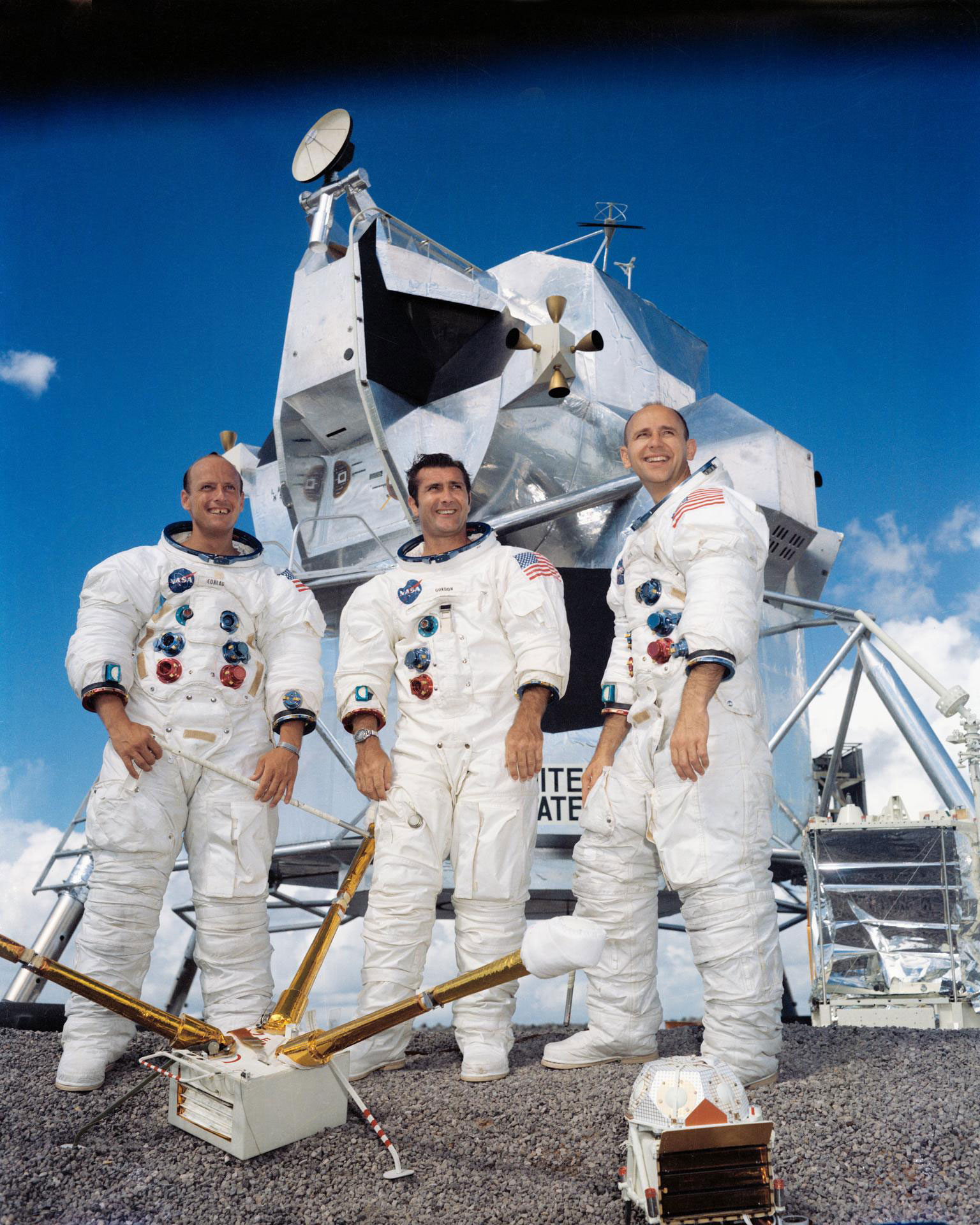 Three men in spacesuits stand in front of a large spacecraft on a sunny day