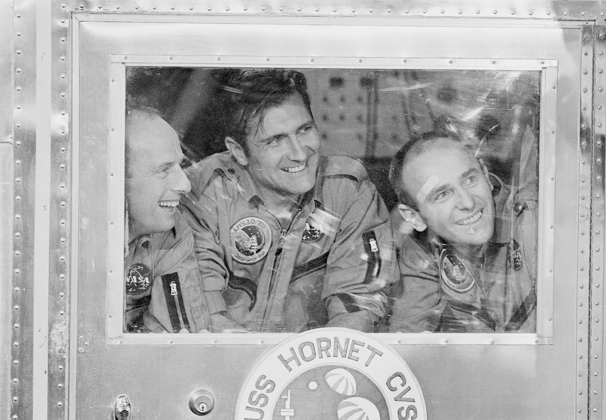 A black and white photograph shows three men smiling from the other side of a ship's window