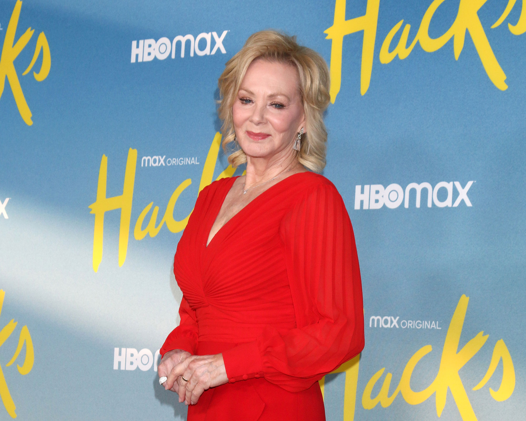 Jean Smart smiling in a red long-sleeved dress in front of a step-and-repeat for the HBO Max show "Hacks."