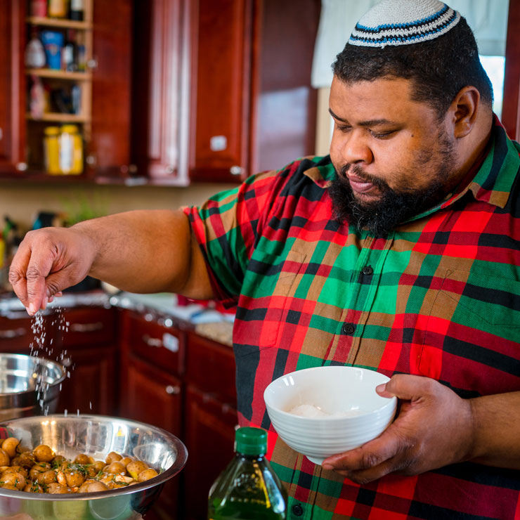 A Black man wearing a yarmulke sprinkles salt on potatoes in the kitchen.