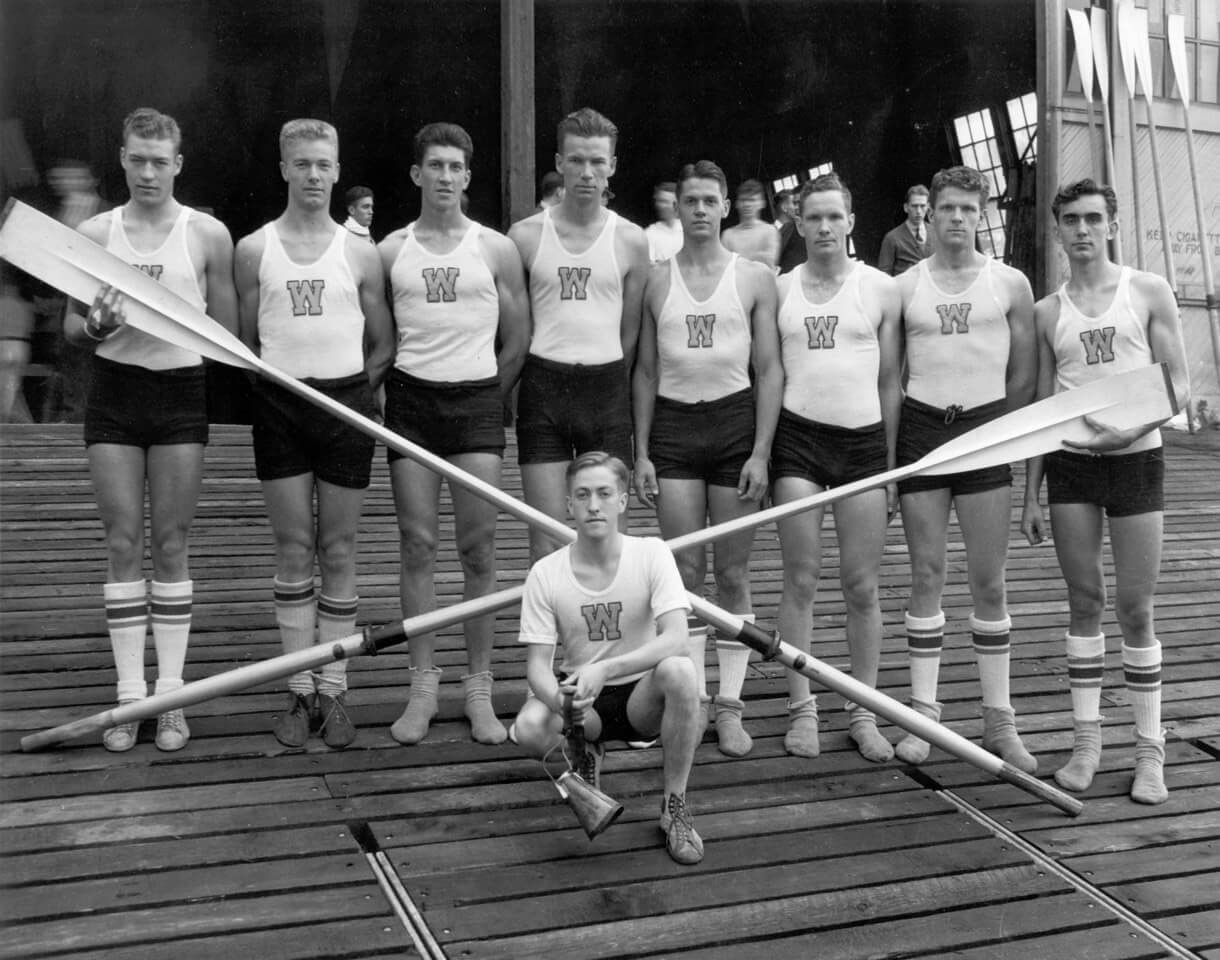 Eight men lined up holding two oars in a crossbar. A ninth man crouches on the ground in front of them.