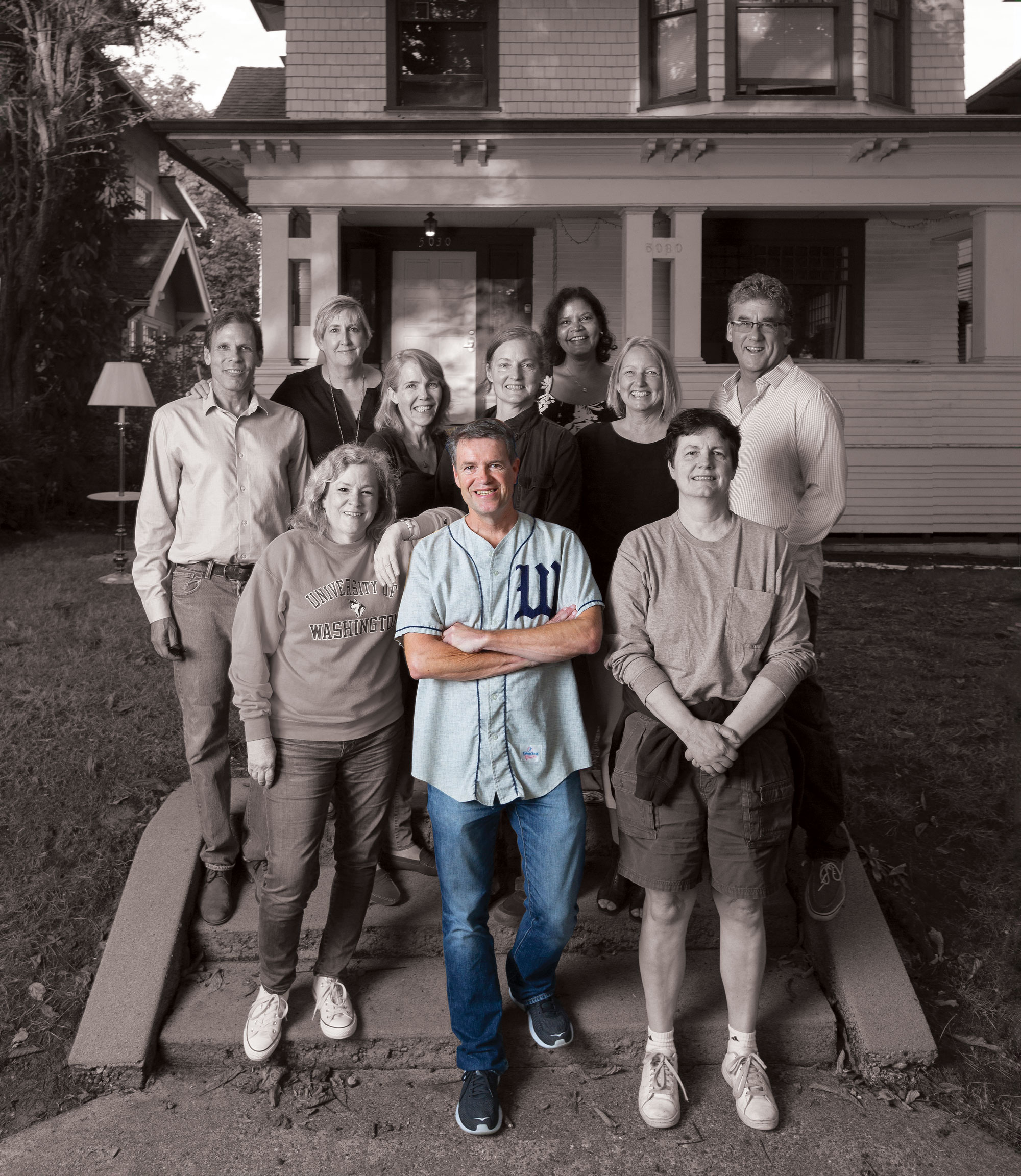 Ten middle-aged people stand in front of a university house smiling. All are in black and white except Jim Caple, front and center.