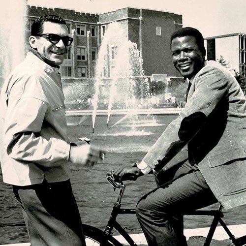 A man wearing sunglasses and a man on a bicycle in front of a large fountain