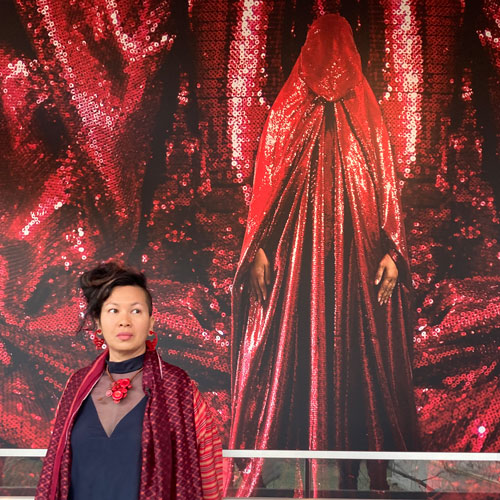 Artist Anida Ali stands in front of a picture of herself wearing a sparkly red chador.