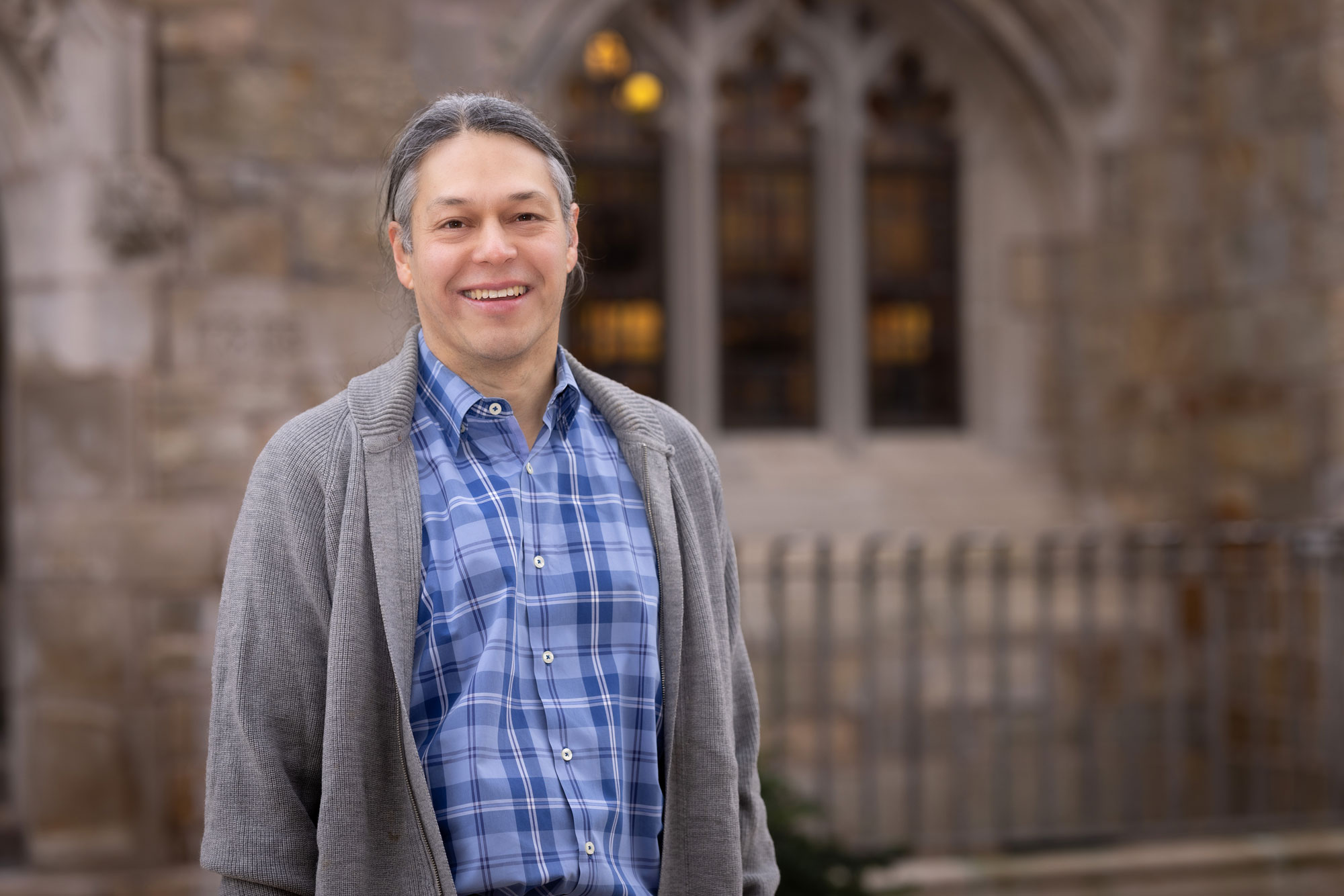 A man in a grey cardigan and blue button down shirt smiles in front of an academic building.