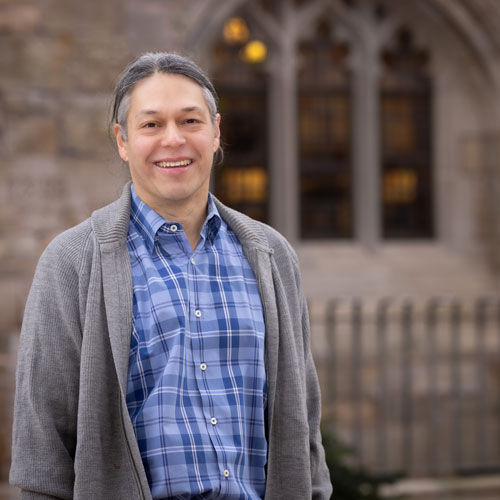 A man in a grey cardigan and blue button down shirt smiles in front of an academic building.