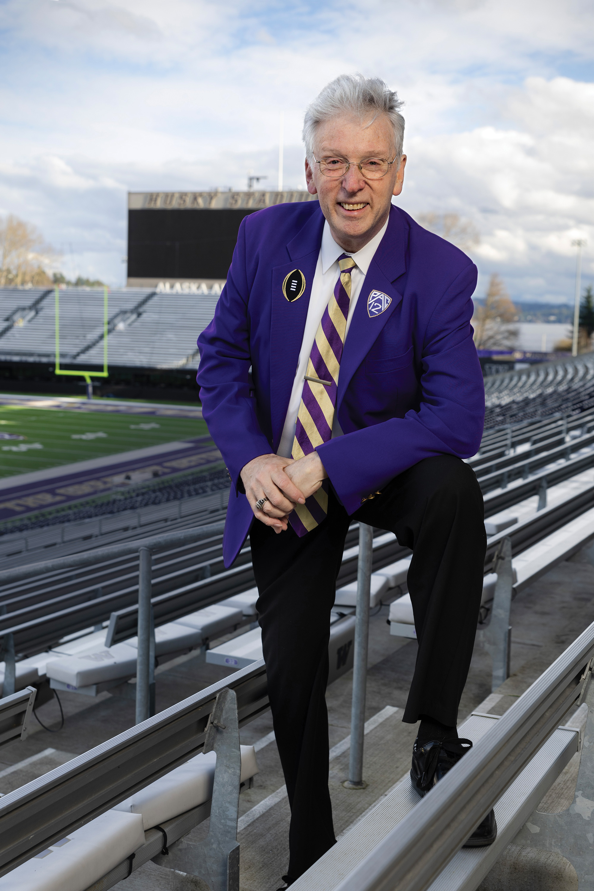 An older man wearing a purple suit and a striped tie smiles in Husky Stadium