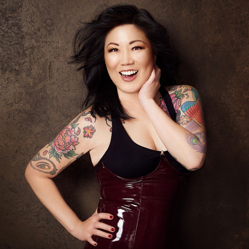 Margaret Cho, a woman wearing a shiny red corset and black tank top, smiles while posing her tattooed arms.