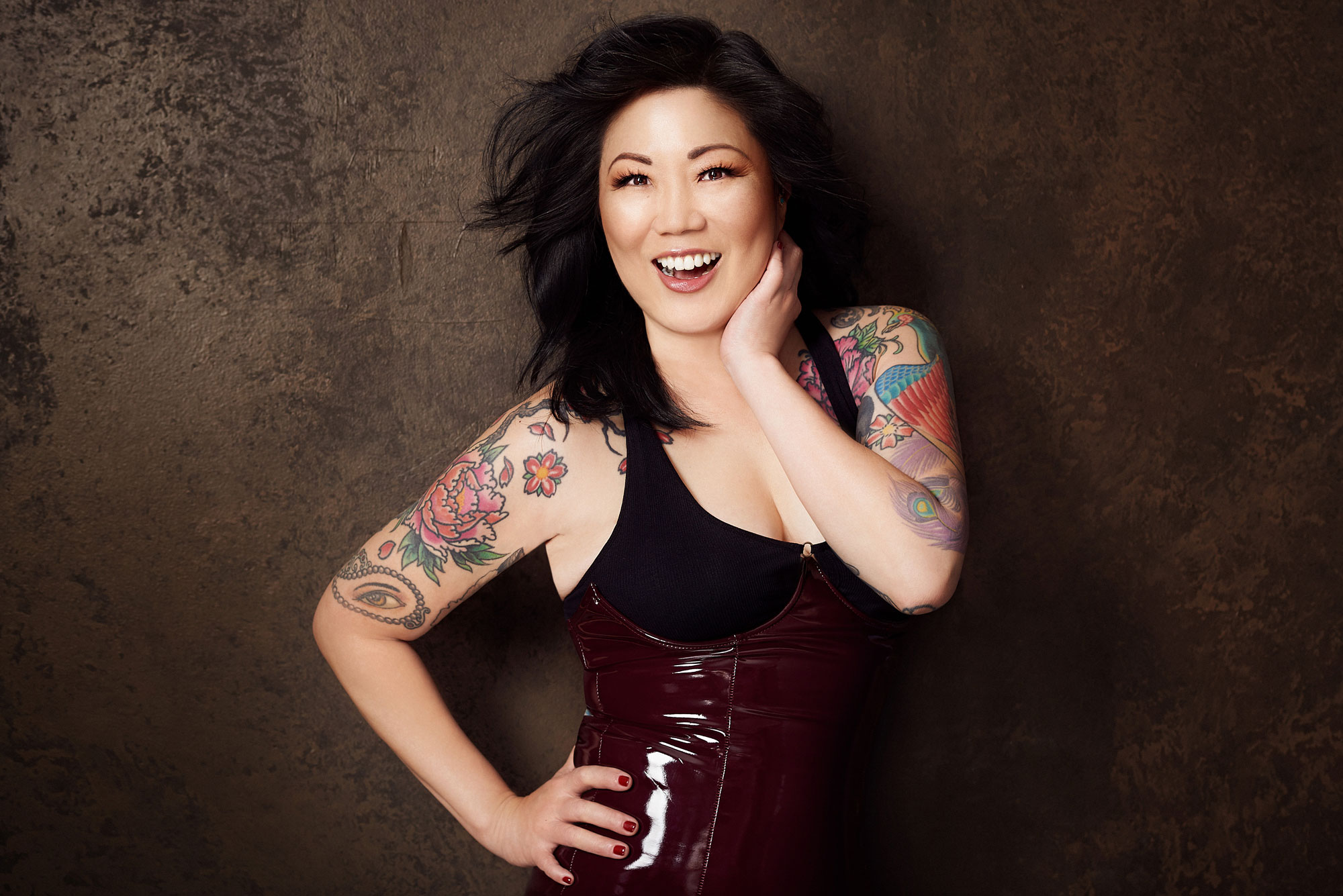 Margaret Cho, a woman wearing a shiny red corset and black tank top, smiles while posing her tattooed arms.
