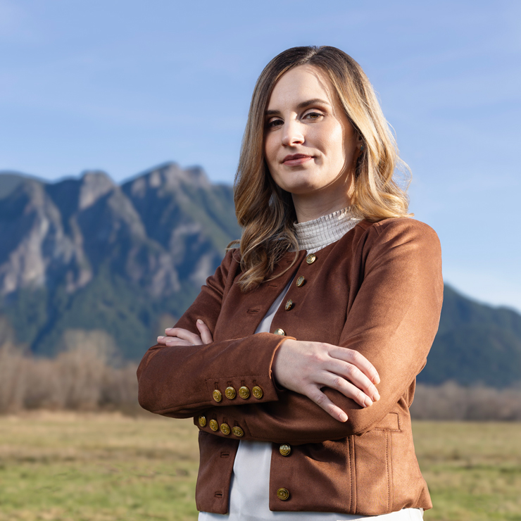 A woman with honey blonde hair, wearing a brown suede jacket, crosses her arms in a large field with mountains in the background
