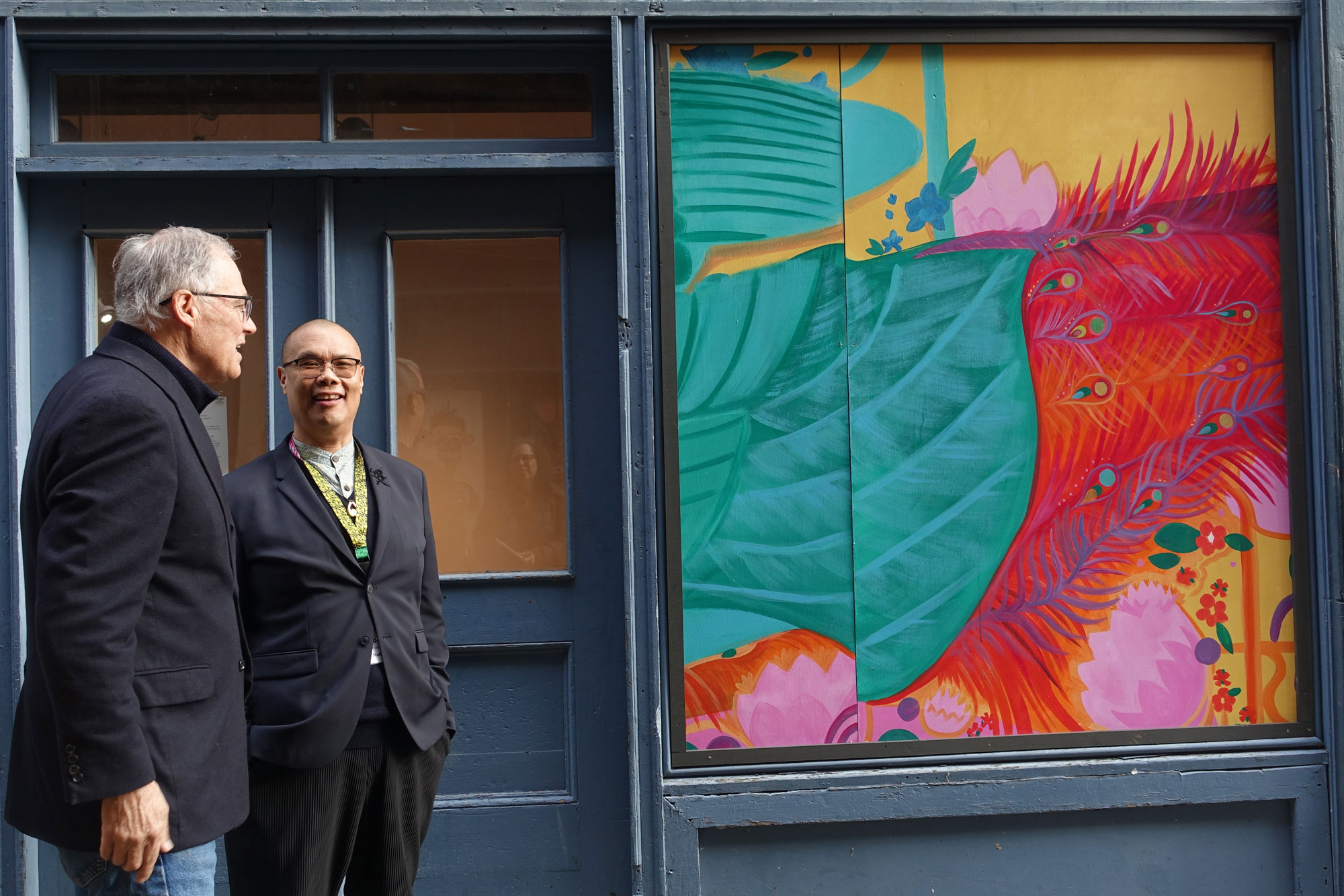 Two men stand in front of a colorful mural in an alleyway