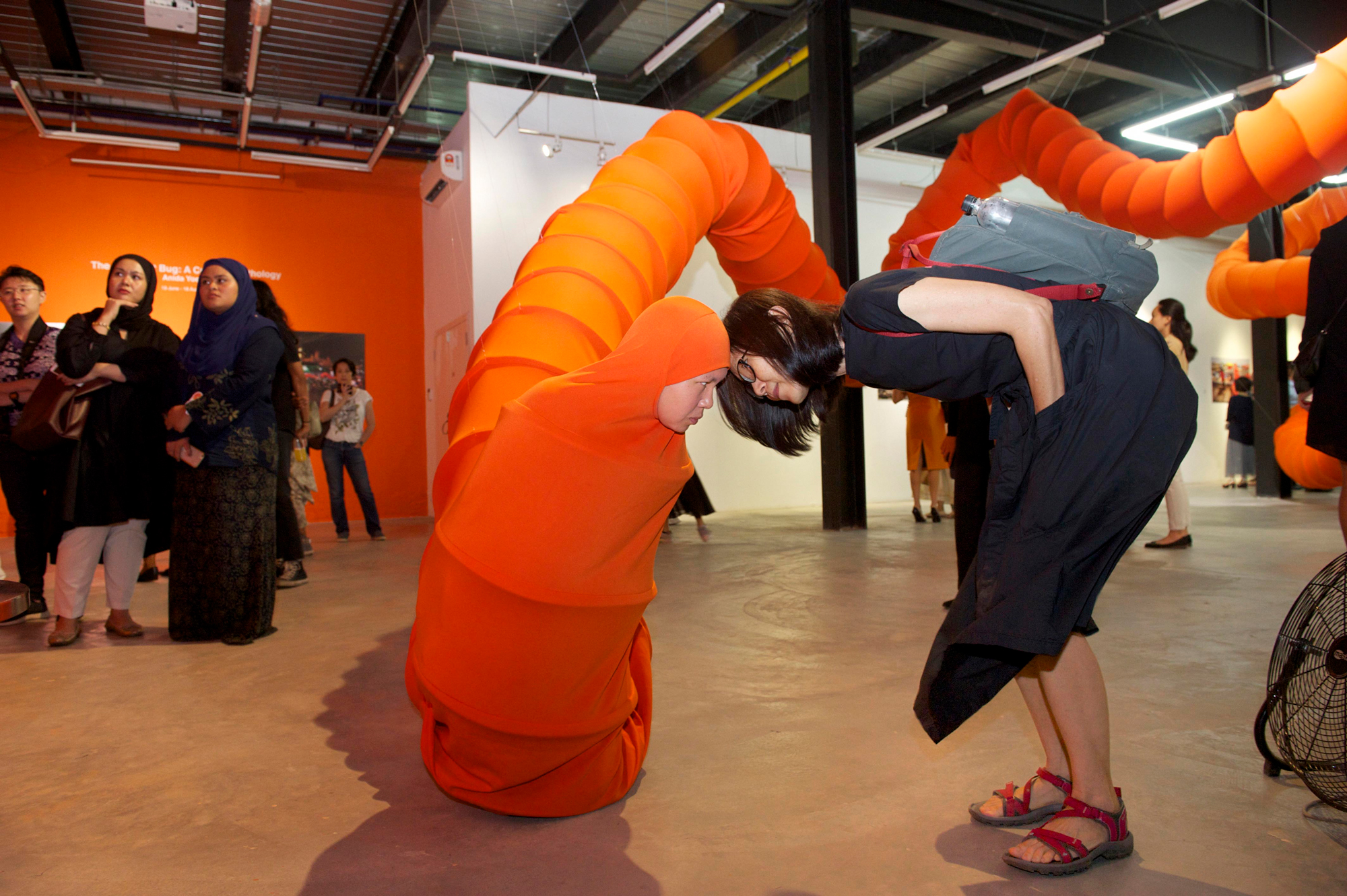 A museum visitor brings her fact close to Anida Ali, who is wearing a giant orange tube that winds around the room