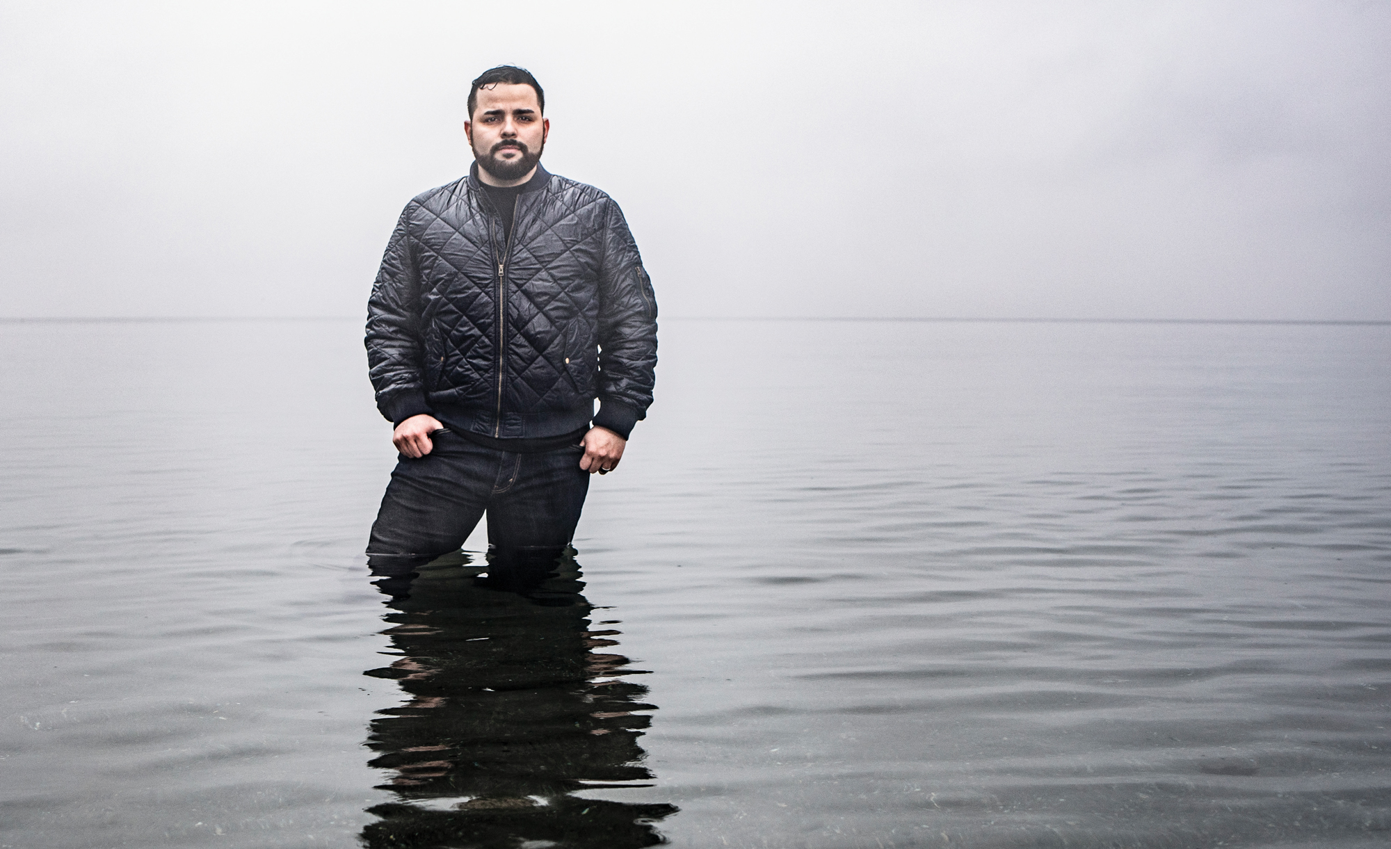 A man with a beard and puffy jacket stands thigh-deep in a body of water on a cloudy day
