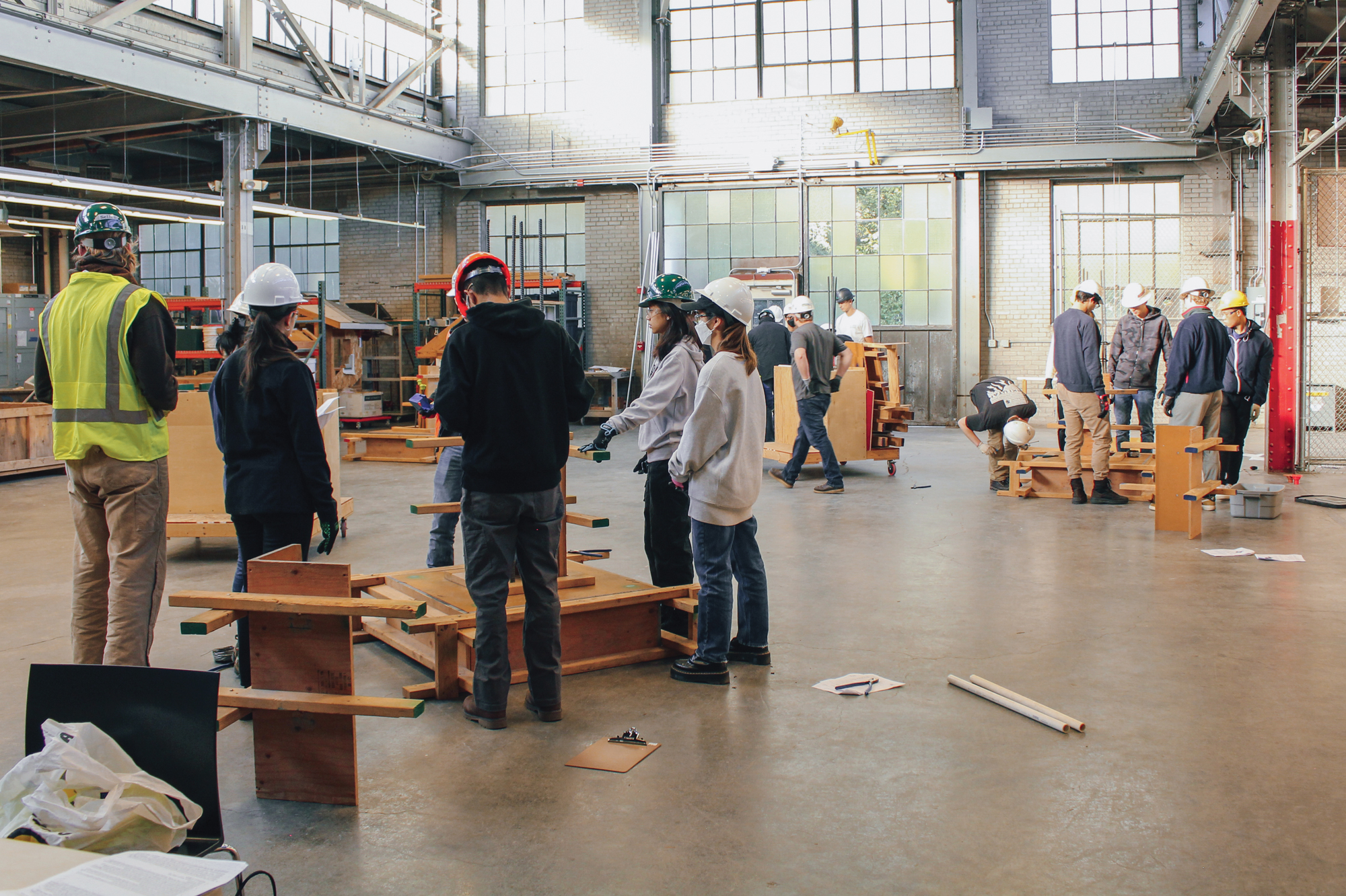 A group of students wearing hard hats and safety goggles in a large room with construction materials