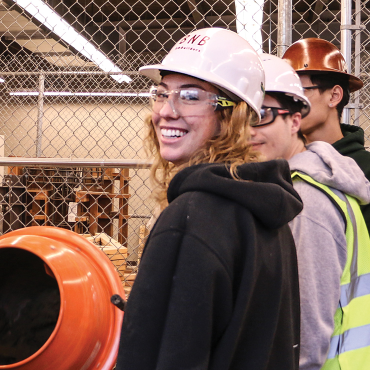 A student wearing a hard hat and safety goggles smiles in front of a cement mixer
