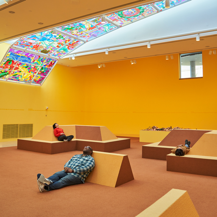 People relaxing in a bright yellow museum room. They're staring at the ceiling, which has a large panel of acetate and tape made to look like stained glass.