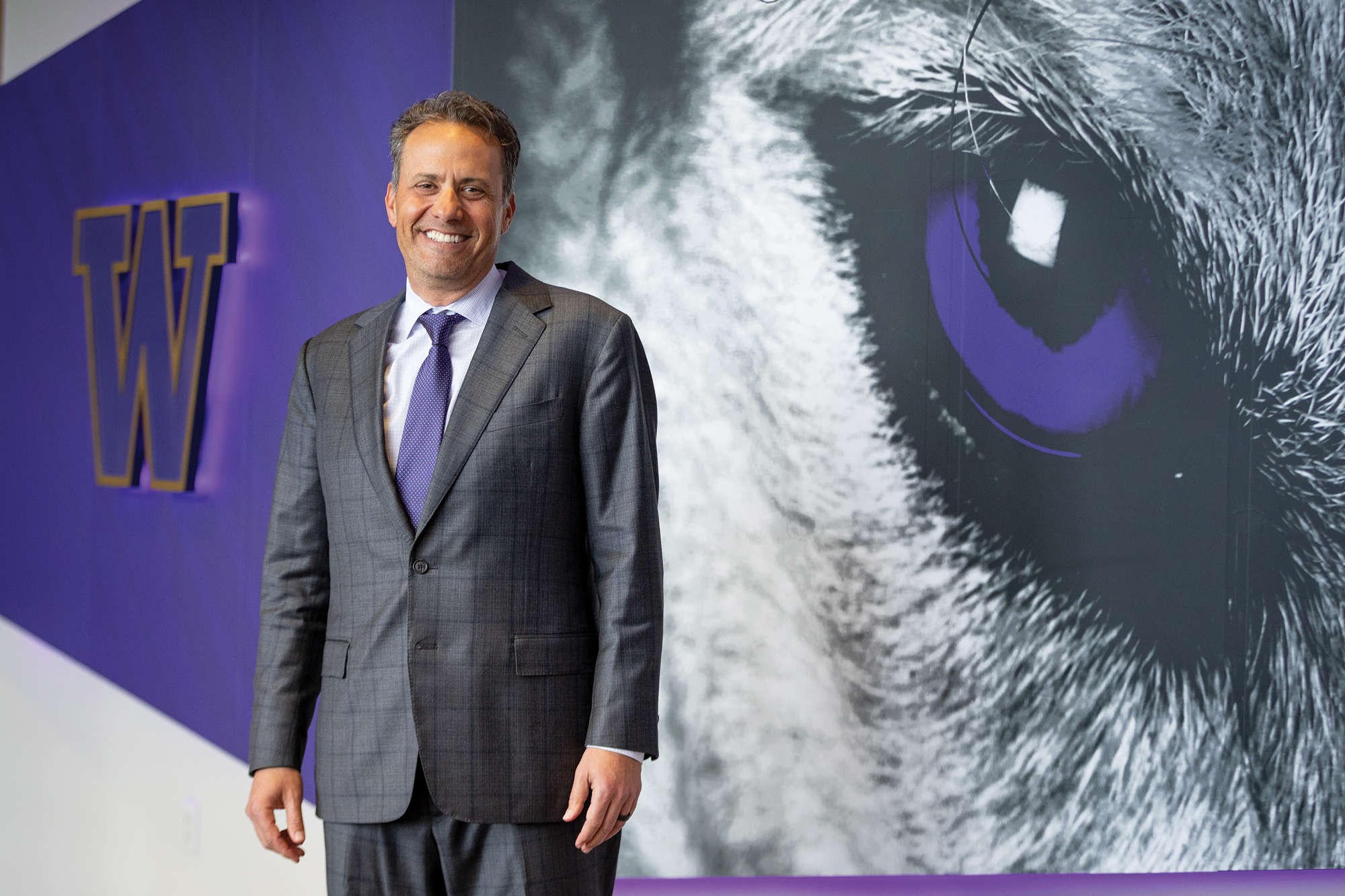 A man smiling, wearing a grey suit and purple polka dot tie, standing in front of a mural of a giant husky