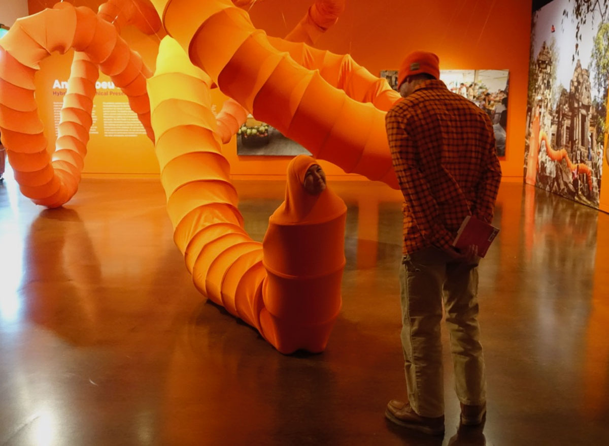 A woman wearing a giant orange tube smiles at an observing man.