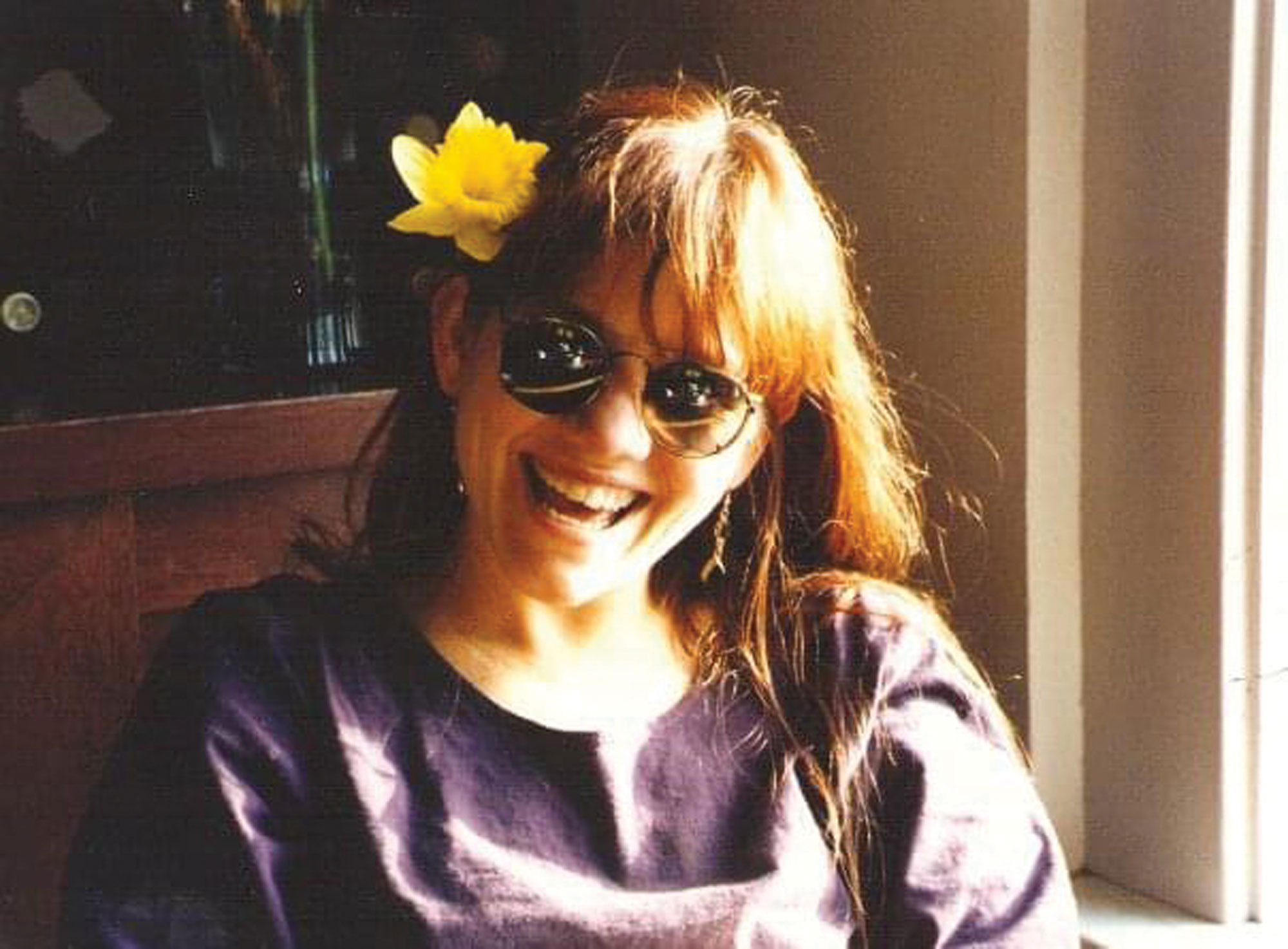 A woman smiling, wearing sunglasses and a yellow flower behind her right ear.