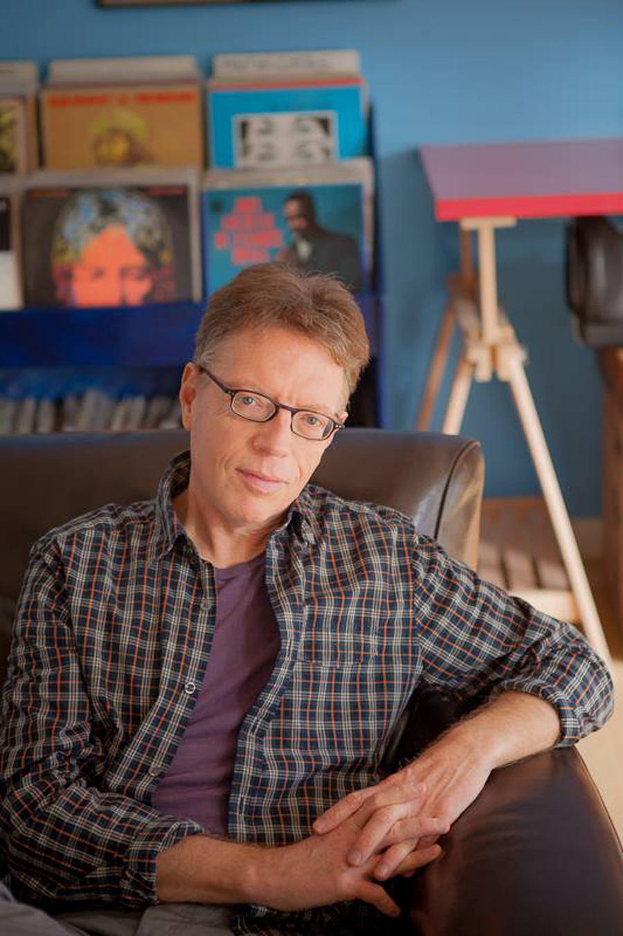 Music journalist Charles R. Cross sits in front of a shelf holding records. He wears a plaid shirt and rests his arm on the couch.