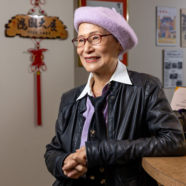 A very stylish woman, wearing a lavender beret, leather jacket, loose tie and lavender blouse - leans against a counter in an office space.