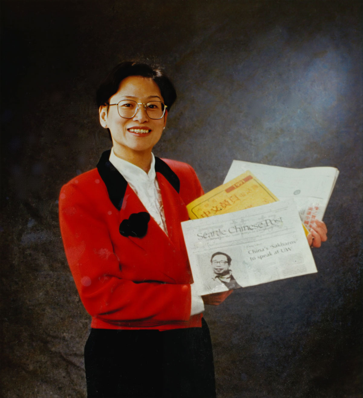 A film photo scan of a young woman in a red blazer and white blouse holding up back issues of Seattle Chinese Post and other Asian American publications.