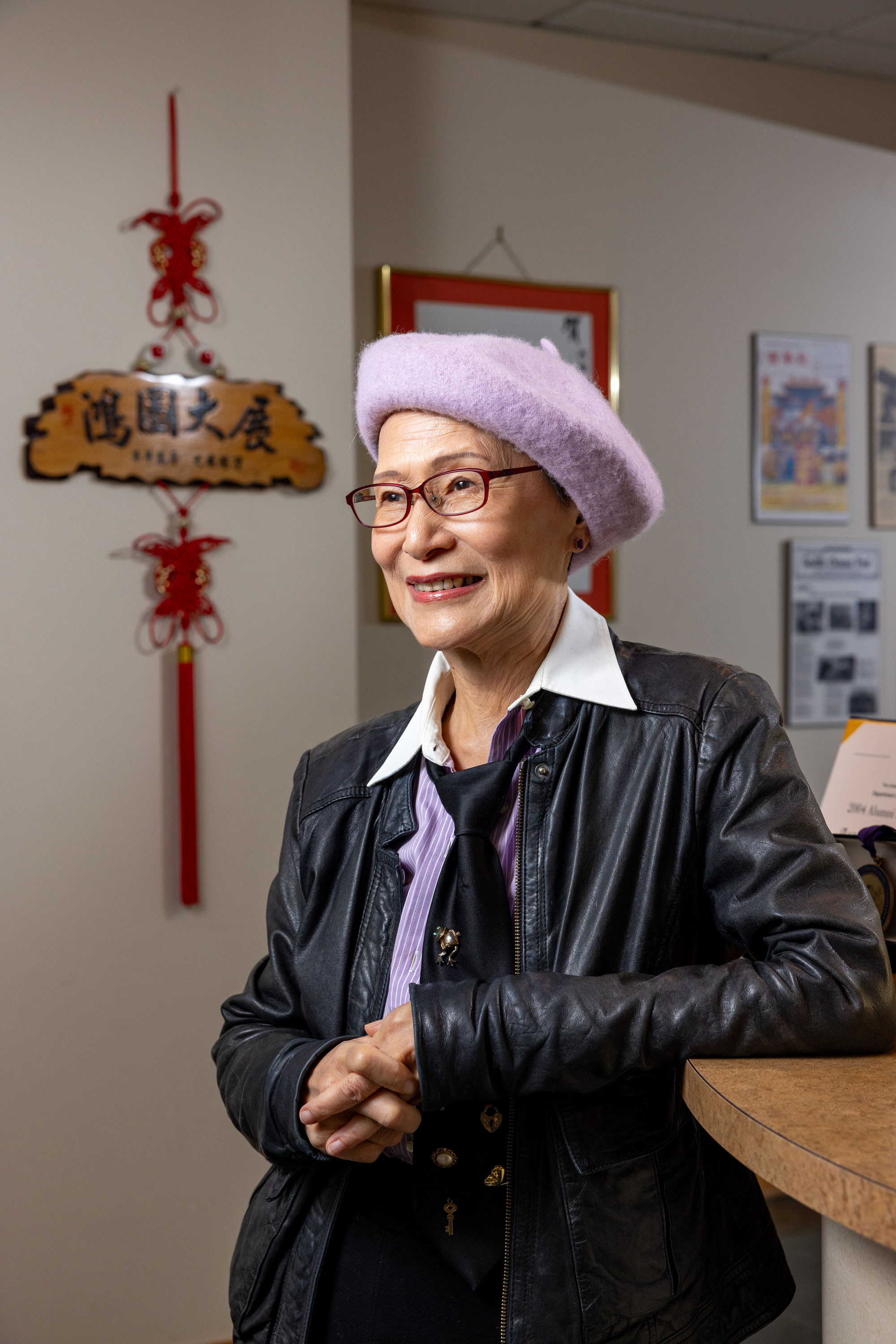 A very stylish woman, wearing a lavender beret, leather jacket, loose tie and lavender blouse - leans against a counter in an office space.