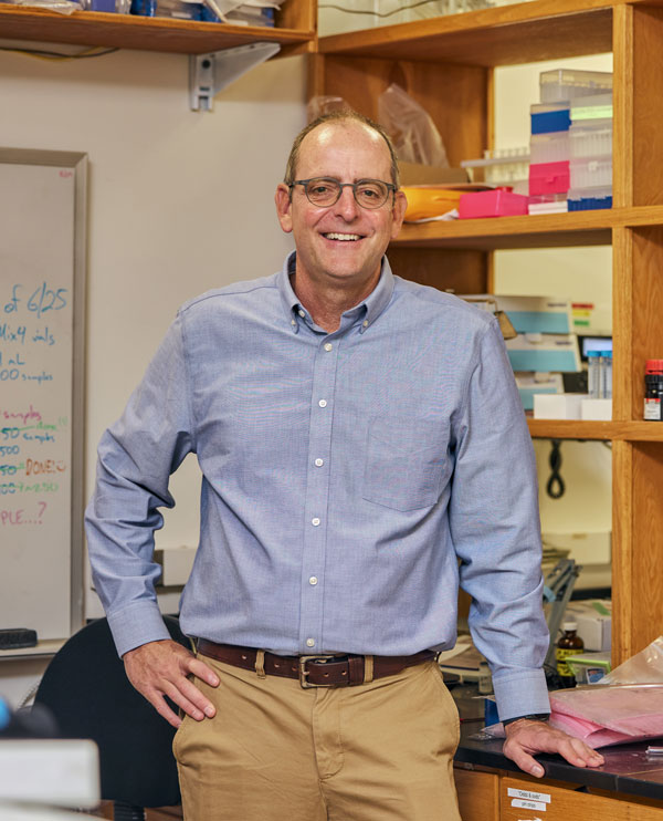 A man in khaki pants and a blue button-up shirt leands on a lab table and smiles.