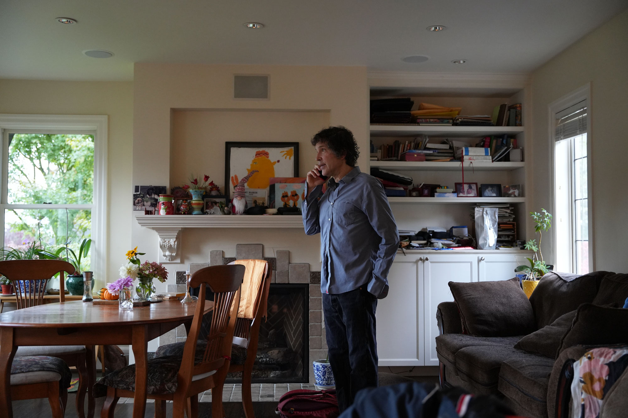 A man in a light blue shirt stands in a sunlit house speaking on the phone.