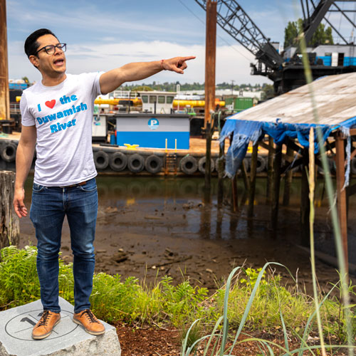 A man wearing a shirt that says "I love the Duwamish River" points to something out of frame in front of the Duwamish River.