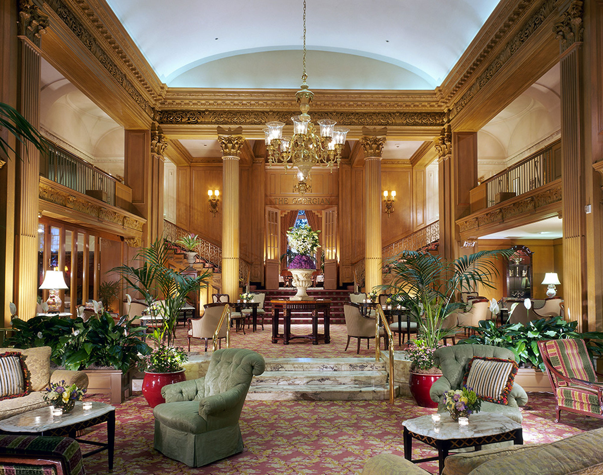 The lobby of an ornate hotel, featuring golden pillars, stuffed armchairs, marble tables and intricately carved moulding.