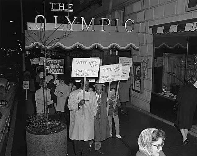 An old photo scan shows a crowd marching in front of the Olympic Hotel with signs that read "Stop Racial Discrimination Now" and "Vote for Open Housing March 10"