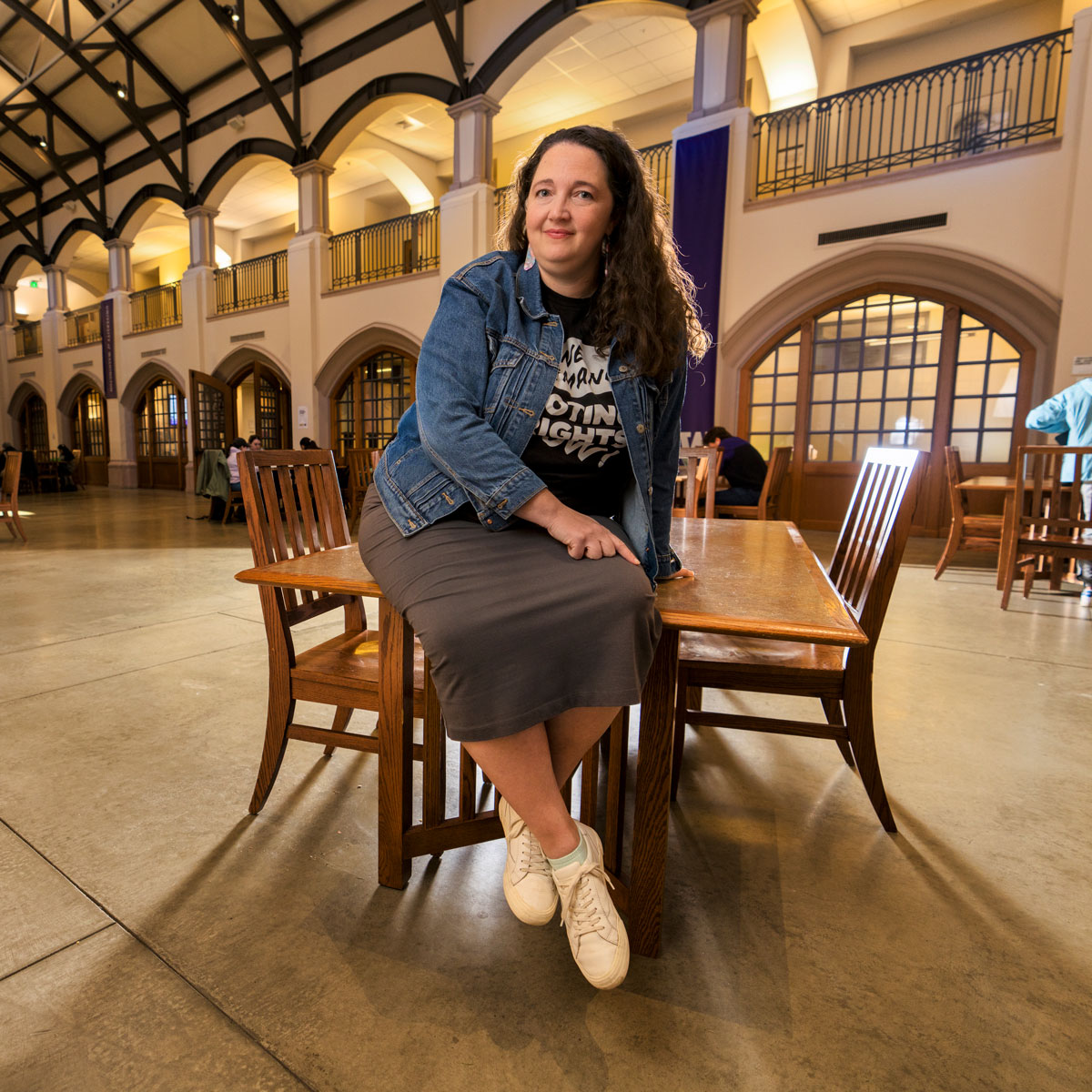 A woman sits on the edge of a table in a bustling student center. She wears a t-shirt that reads "We Demand Voting Rights Now."