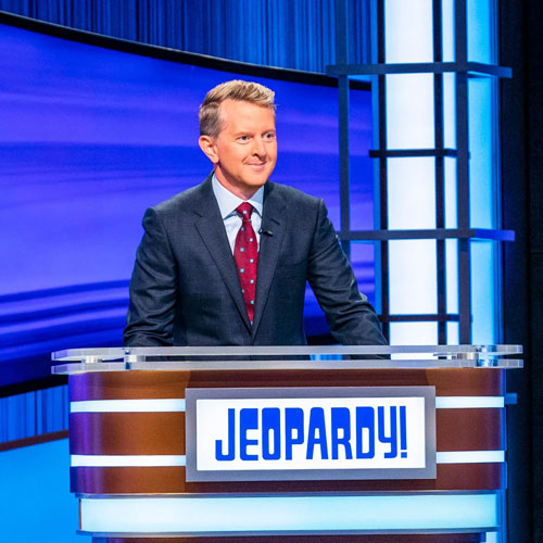 Ken Jennings wearing a suit, standing behind a podium that reads "Jeopardy!"