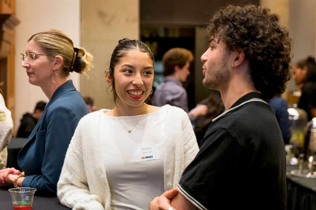 A woman wearing white smiles while speaking to a man in a black polo shirt at a reception event.