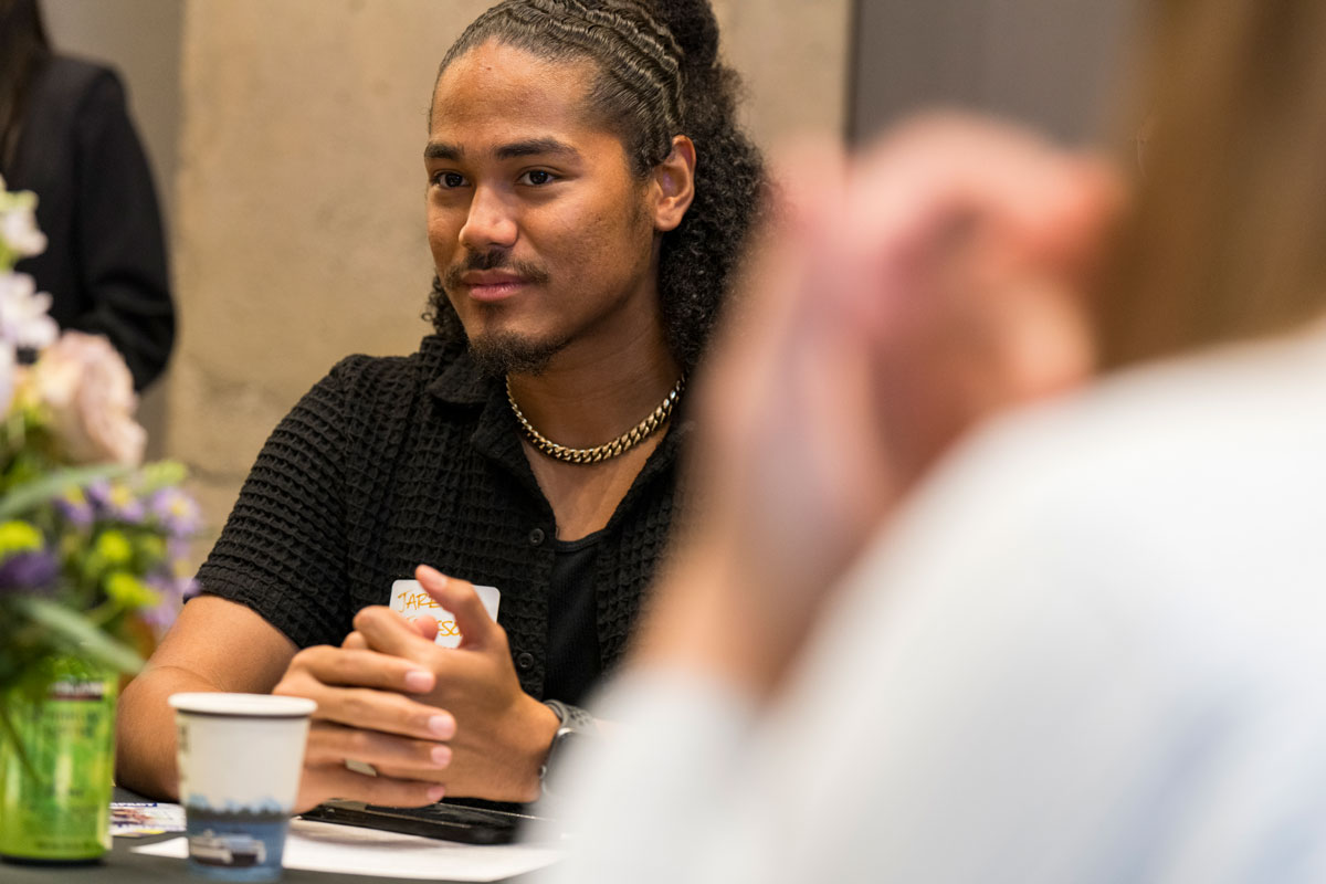 A man wearing long hair, a gold necklace and a black shirt sits at a formal table, smiling while listening to a speaker out of frame.