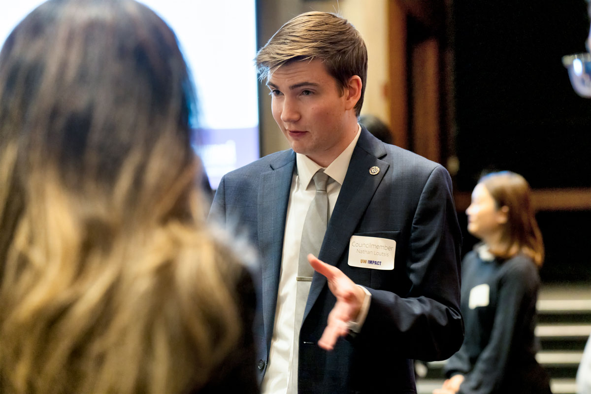 A man with blonde hair and a blue suit speaks to a woman whose face is turned away from the camera. His nametag reads "Councilmember Nathan Loutsis."