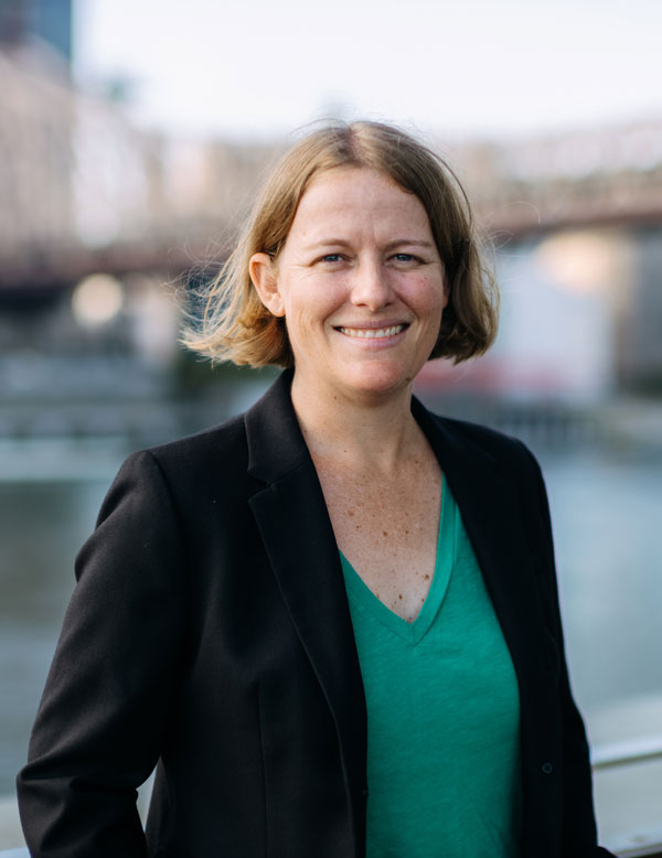 A woman in a green blouse and black blazer with a blonde bob smiles in front of a body of water in an urban area.