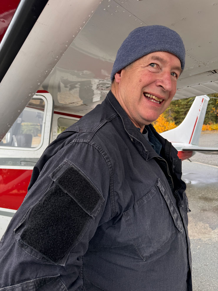 A man wearing a flight jacket and knit beanie smiles outside of a small airplane.