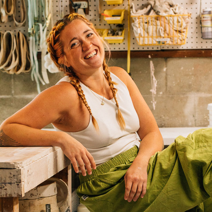 A woman in a white tank top and green pants with braided hair leans against a workbench and smiles