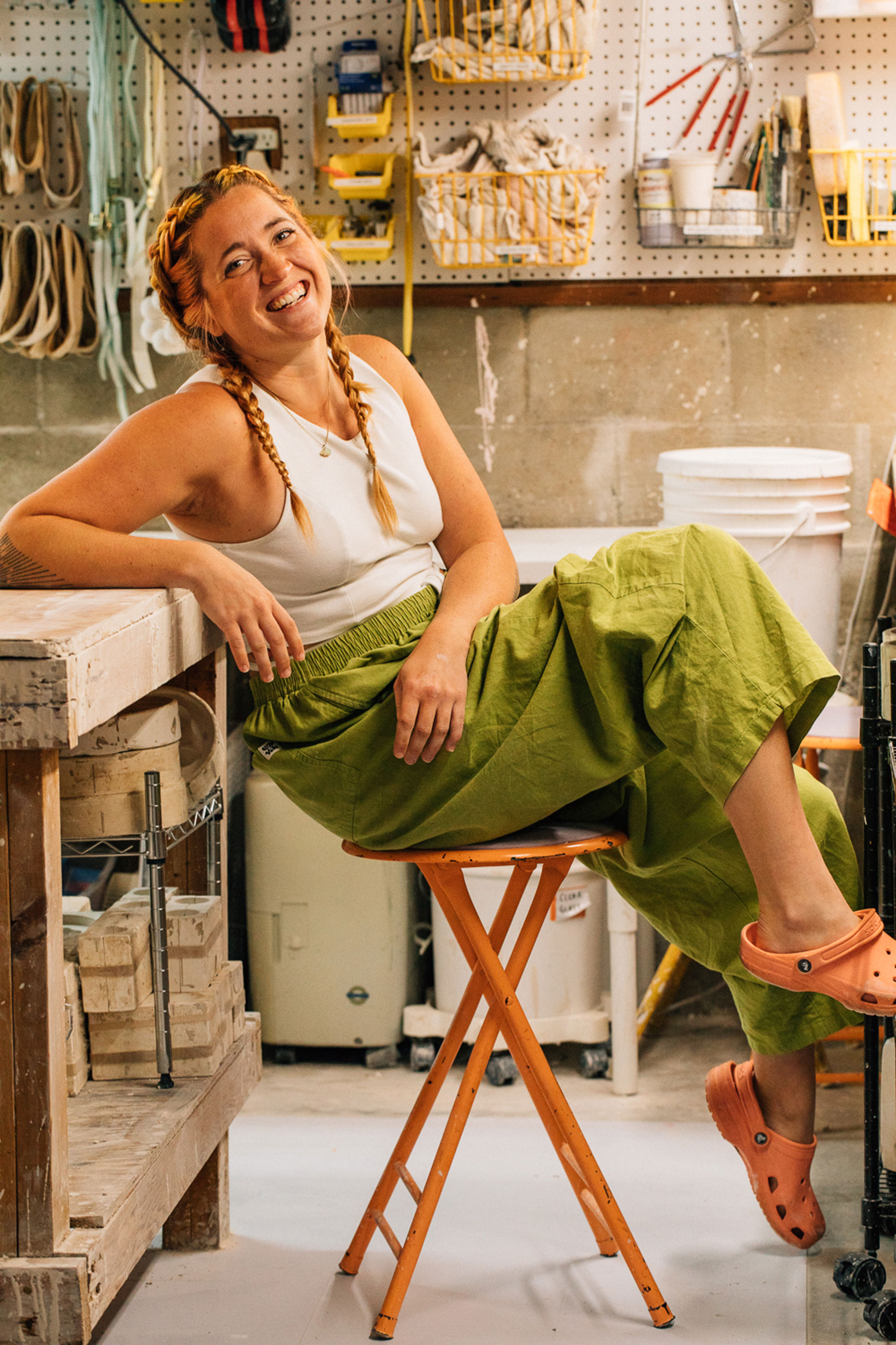 A woman in a white tank top and green pants with braided hair leans against a workbench and smiles