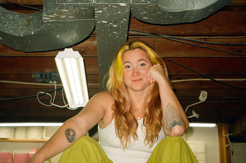 A woman with dyed yellow hair and geometric tattoos sits in her art studio